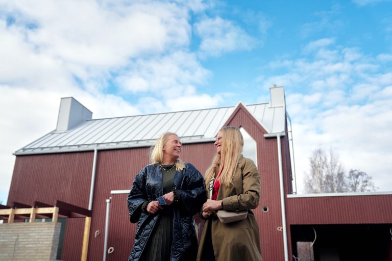 Hanna Luukkonen (vas.) ja Riikka Säkkinen katsovat toisiaan hymyillen punaisen talon edessä.
